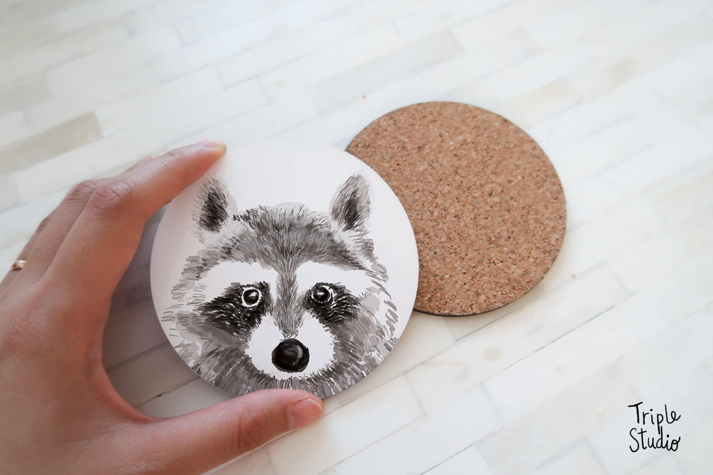 May include: Two round coasters. One coaster features a detailed black and white illustration of a raccoon's face. The other coaster is made of cork. The coasters are designed for use with beverages and to protect surfaces.