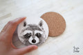May include: Two round coasters. One coaster features a detailed black and white illustration of a raccoon's face. The other coaster is made of cork. The coasters are designed for use with beverages and to protect surfaces.