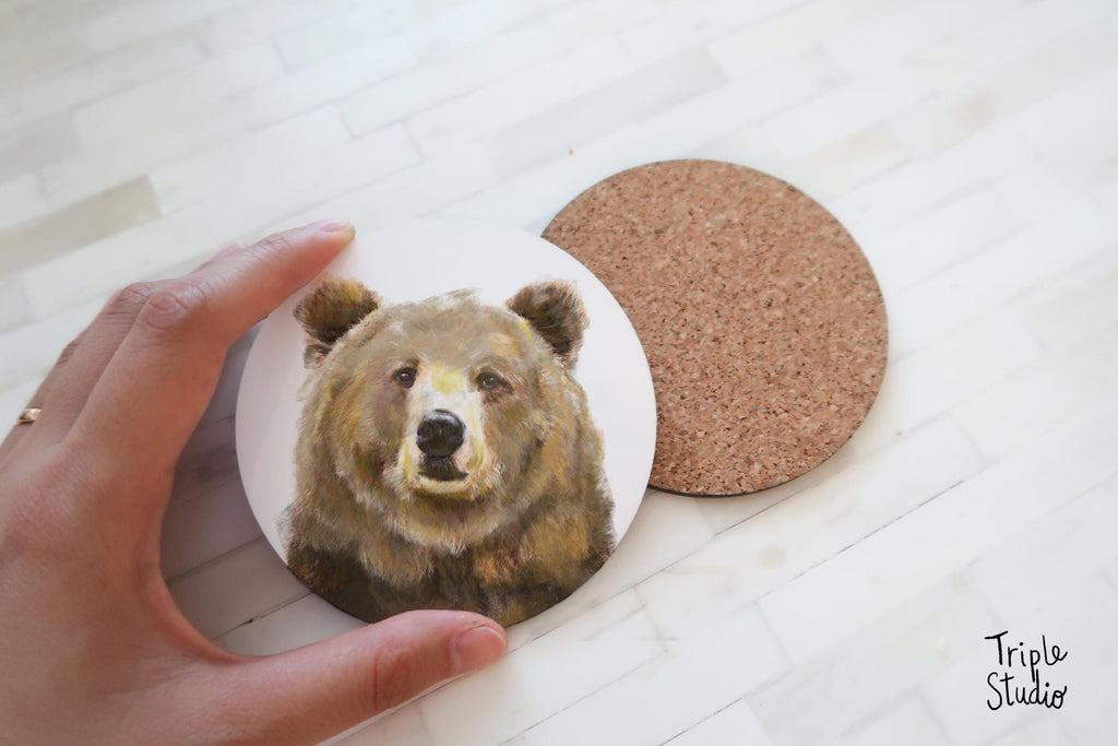 May include: Round coaster with a detailed painting of a brown bear's face. The coaster is white with the bear image, and a separate cork coaster is next to it. The image is from Triple Studio.